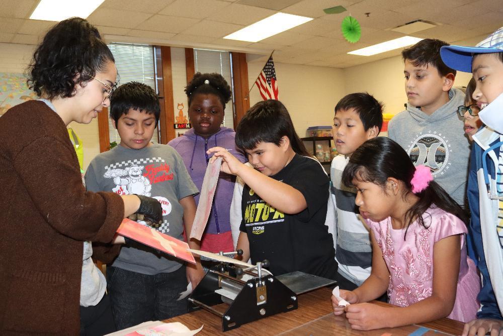 There are nine people in a row in the photo. At far left, guest artist Angelica Tapia prepares to take part in a printmaking activity with Everett Elementary School students.