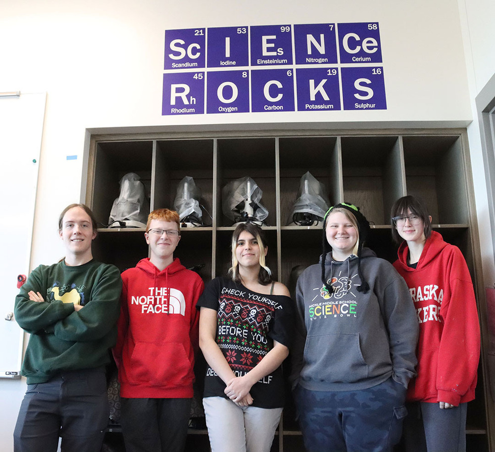Five Science Focus Program students stand in a classroom in a row. They are standing in front of a cubbyhole shelf that is holding many microscopes. A sign that reads Science Rocks is above the cubbyholes. There are two boys and three girls in the photo.
