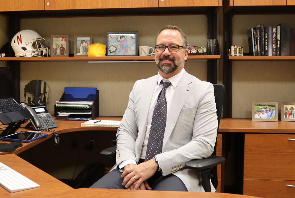 LPS Director of Security Nate Hill smiles while sitting in a chair in his office. He is sitting at his desk in front of a shelf. The shelf contains pictures, books and artwork from his family. There is a Nebraska football helmet on the lefthand side of the shelf.