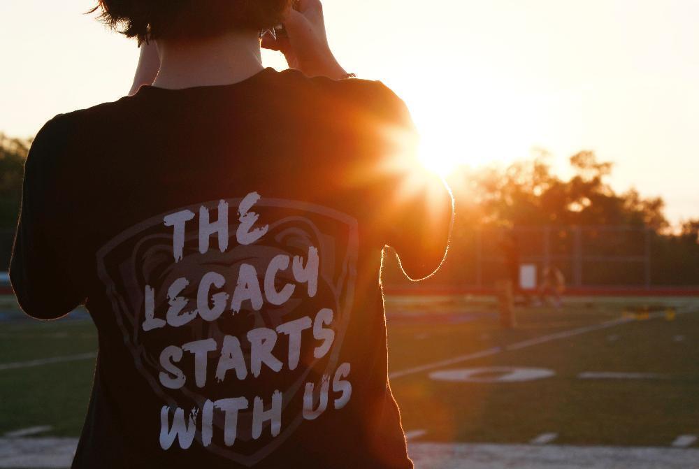 A person is pointing a smartphone toward the rising sun while wearing a t-shirt that says The Legacy Lives at Grizzly Senior Sunrise.