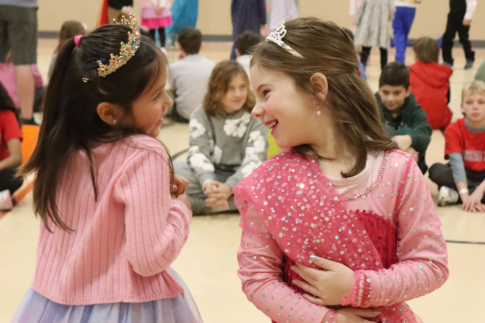 Two kindergarten students smile at each other as they take part in a royal ball in the gym. Both are wearing tiaras on their heads.