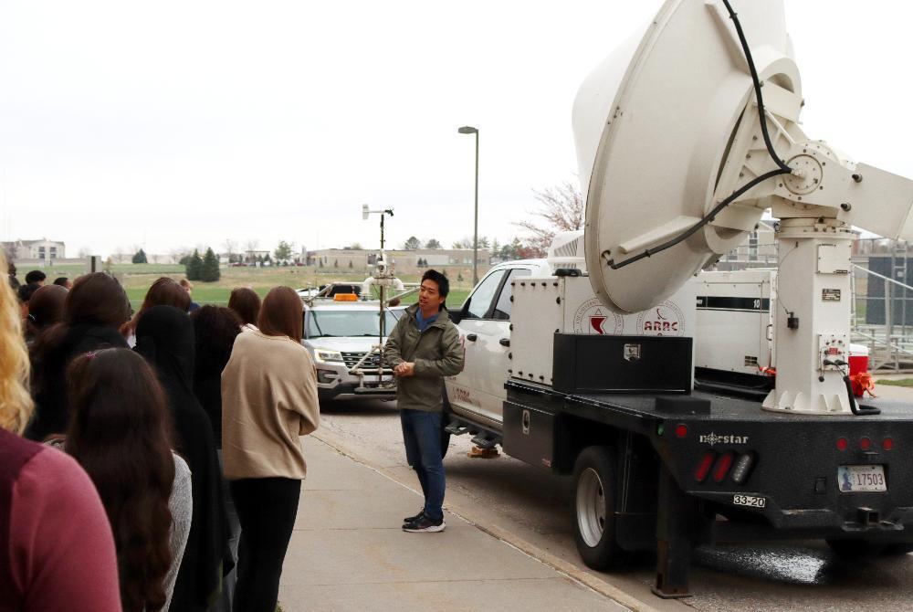 A man explains a large satellite dish mounted on a truck to a group of students outside. The license plate reads "L 17503".