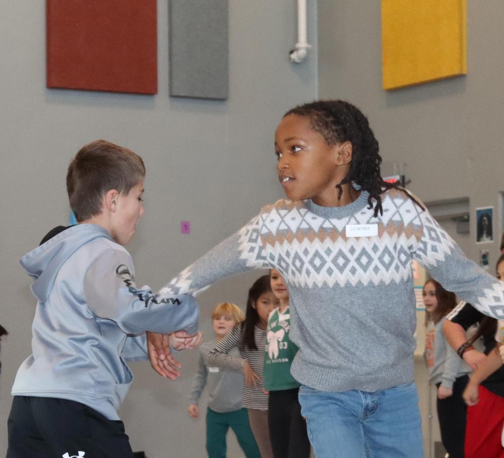 Robinson Elementary School third graders smile as they take part in dancing lessons during their Backstage at the Ballet experience. A girl and boy are holding hands as they twirl in a circle during a partner portion of their dance. The girl is wearing jeans and a sweater, and the boy is wearing windpants and a long-sleeved workout shirt.