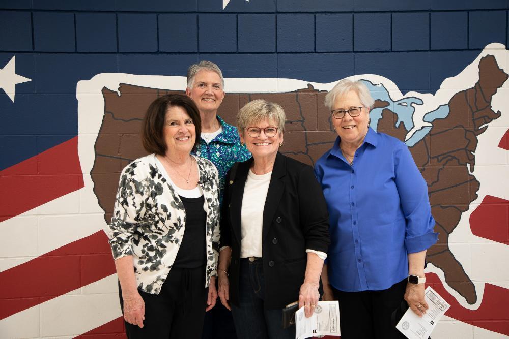 Four smiling older women stand together in front of a colorful mural of the USA map and an American flag. They are casually dressed, and two women in front hold papers.