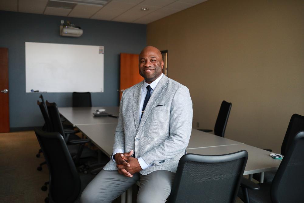 A smiling man in a light gray suit sits on the edge of a conference table in a meeting room with chairs, a whiteboard and a projector behind him.