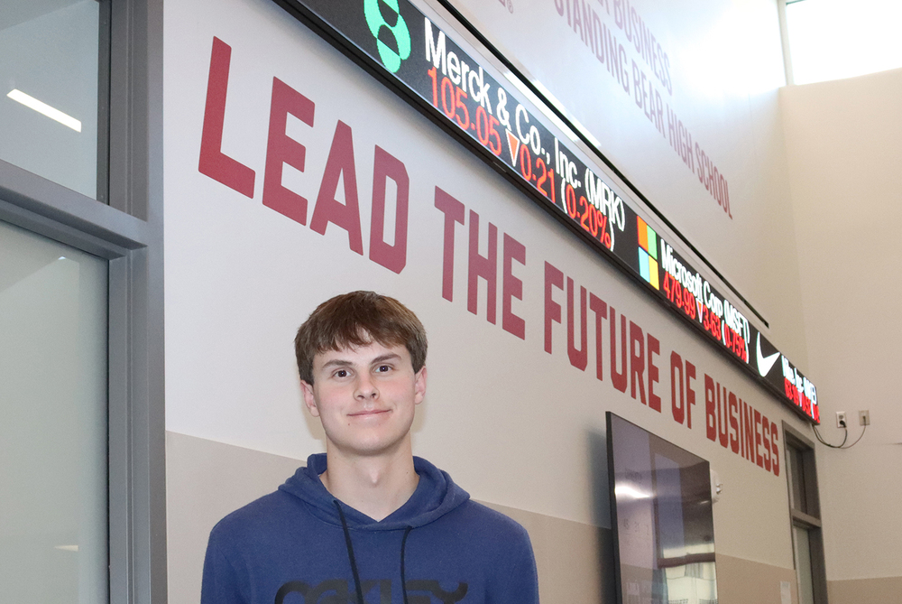 Nebraska Business Focus Program senior Ashton stands outside the main classroom at Standing Bear High School. The phrase 'Lead the Future of Business' is painted on a wall underneath an electronic stock market ticker.