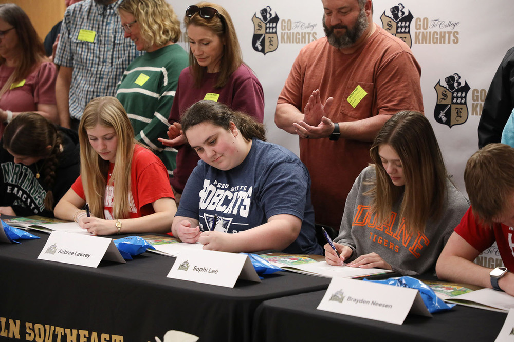 Five Southeast High School seniors use pens to sign their names in books during a signing ceremony for future educators. Several adults are standing behind them and clapping.