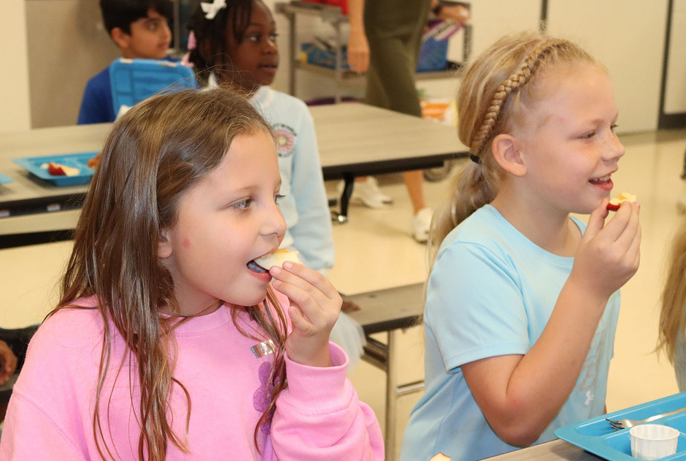 Two Maxey Elementary School students smile as they participate in Apple Crunch Day. The girl on the left is taking a bite of an apple slice while holding the slice with her left hand. The girl on the right is holding an apple slice in her right hand and is preparing to take a bite of it.
