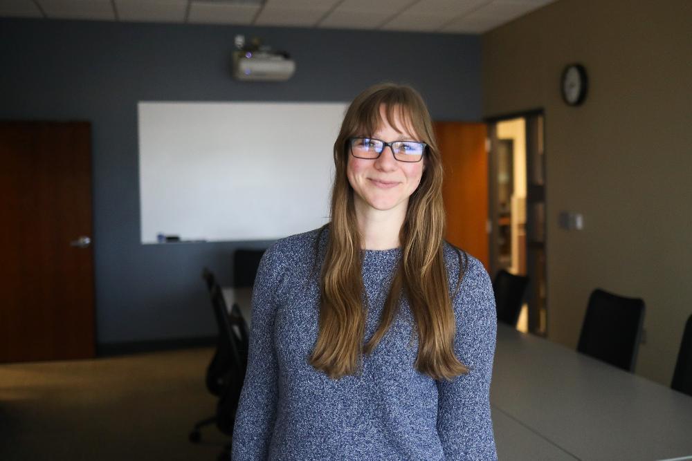 A young woman with long brown hair and glasses smiles in a meeting room. She wears a blue patterned sweater. Behind her are a whiteboard, projector, and conference table with chairs, set against a two-toned wall.