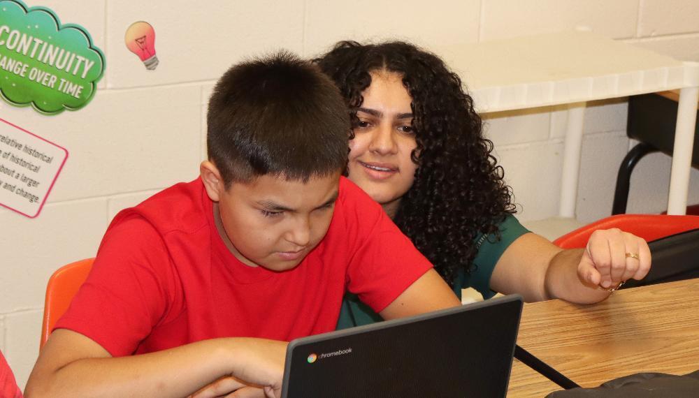 Goodrich Middle School social studies teacher Hilan Younis speaks with a seventh grade student as he conducts research for a class assignment. The student is sitting at a desk and looking at his laptop computer. Hilan is crouching down next to him and is smiling as she looks at his research.