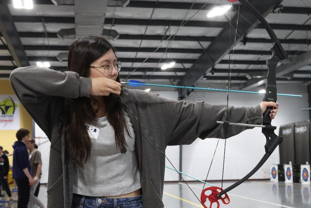 A young woman with long dark hair and glasses aims a black recurve bow with a blue arrow, drawing it back to her cheek. She wears a grey cardigan and stands in an indoor archery range with targets visible behind her.