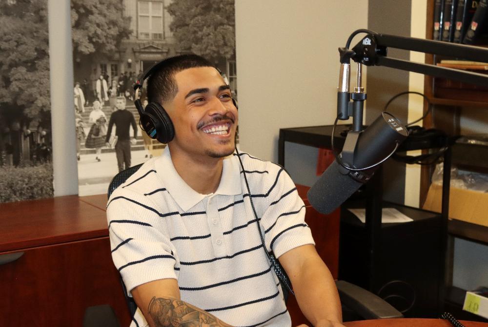A young man with headphones and a striped shirt smiles broadly, looking left, seated at a desk. A studio microphone hangs before him. Behind him is a vintage black-and-white image of a building and people.