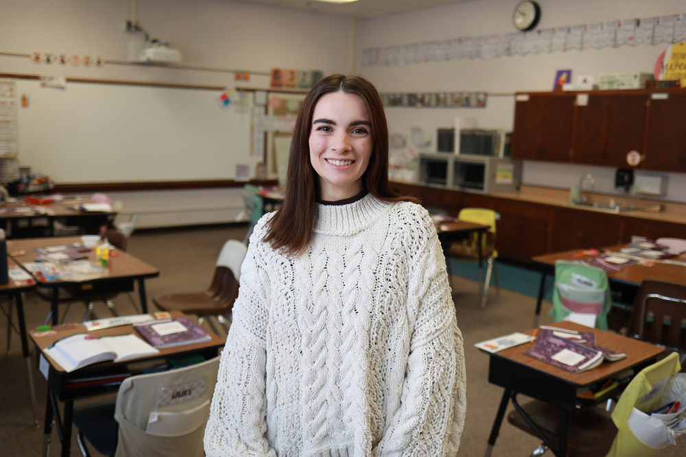 Isabelle Short Gomez smiles in her classroom at Randolph Elementary School. There are several rows of desks behind her that have books and school supplies on them. There are several bookshelves and a large whiteboard in the background.
