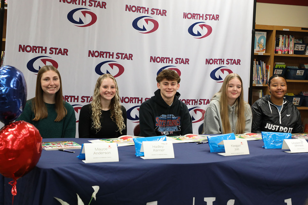 Five North Star students smile as they sit together in a row at a table during a signing ceremony. There are books in front of them and a banner with the North Star logo behind them.