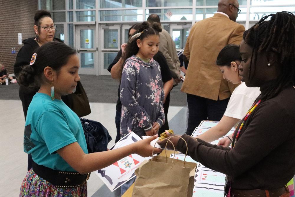 A girl receives a paper bag from a woman. The exchange happens at a table with papers and other people nearby in a large hall. The paper shows the text: "Indigenous Education Council".