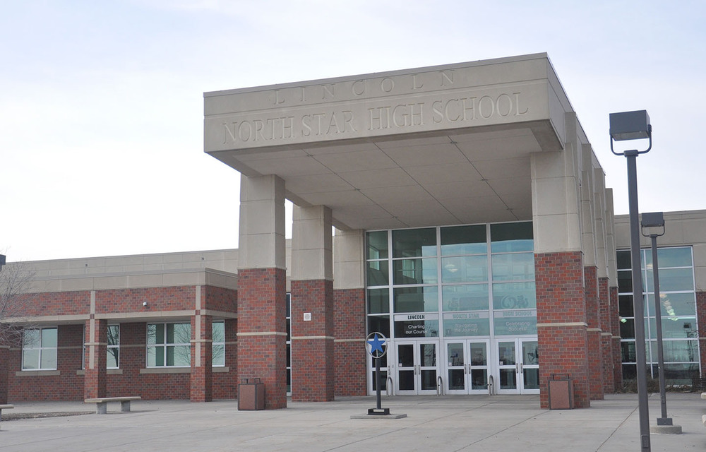 The front entrance of Lincoln North Star High School is shown. Six pillars are holding up a large outdoor ceiling with the words 'Lincoln North Star High School' engraved on it. There are four sets of double doors at the ground level and a wall full of windows above the front entrance.