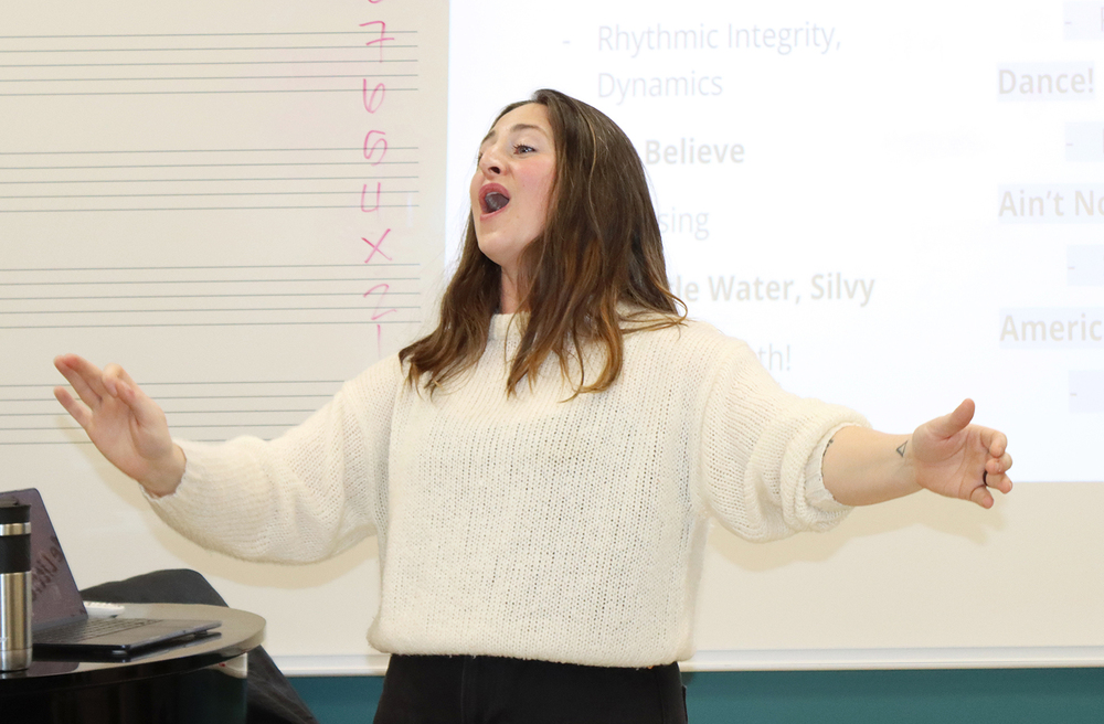 Moore Middle School music teacher Hannah Lambert leads a class at the front of her room. She is waving both hands in the air while her choir sings a song.