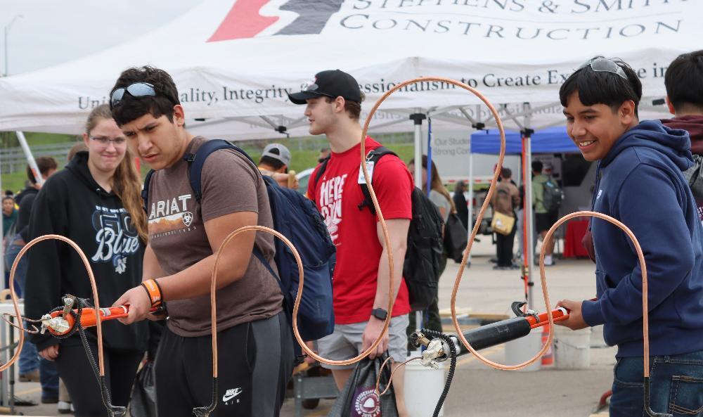 Two young men manipulate flexible copper tubing connected to hand tools at an outdoor event. The event features tents and a banner reading "Stephens & Smith CONSTRUCTION," "Unity, Integrity, Professionalism to Create Excellence."
