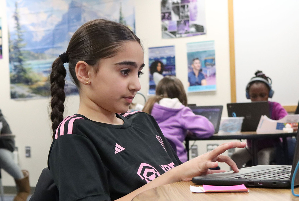 A Lakeview Elementary School student sits at her desk in a computer science classroom. She is looking at a laptop and moving the cursor with her right hand on the keyboard.