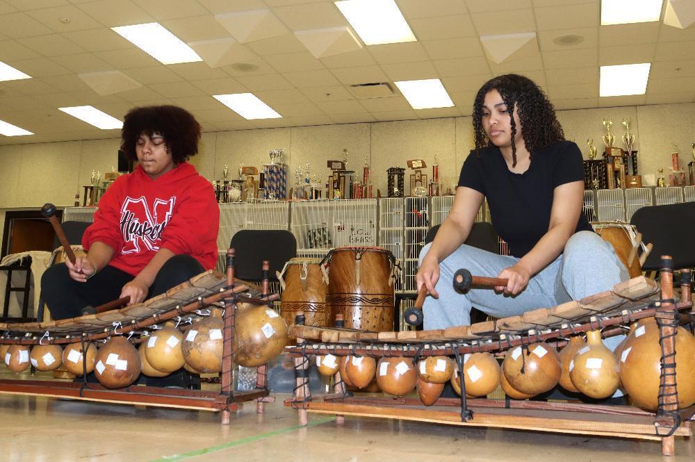 Two students sit, playing large wooden xylophones with gourd resonators, holding mallets. They are seated in a room with shelves of trophies and drums behind them.