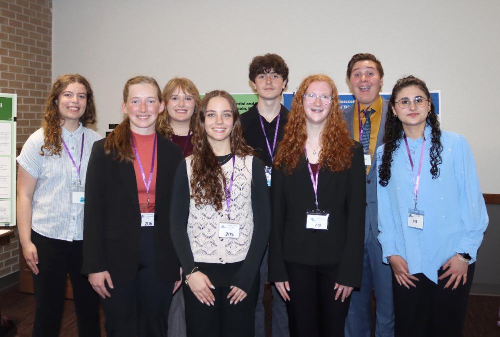 Eight smiling young adults—seven women and one man—pose for a group photo. They wear purple lanyards and ID badges in front of presentation posters, likely at an academic event.