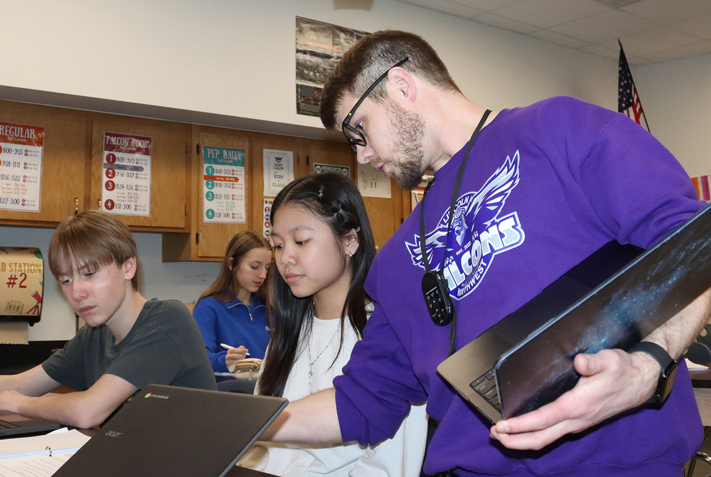 Northwest High School teacher Anton Olbricht looks over a student’s analysis of a thermoregulation science experiment in class. He is wearing a long-sleeved shirt that has the Northwest Falcons logo on it. The student is sitting at her desk and a laptop computer is in front of her.