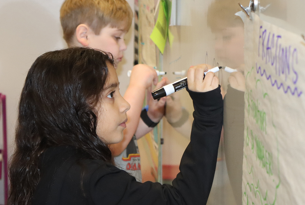 A Zeman Elementary School student writes on a whiteboard by holding a marker in her right hand. She is standing next to a classmate by the board.