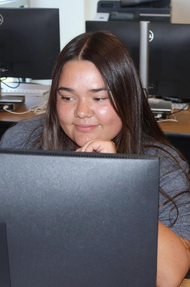 A young woman with long dark hair and freckles smiles softly, resting her chin on her hand. She is intently looking at a computer screen, with other monitors visible in the background, suggesting a computer lab setting.