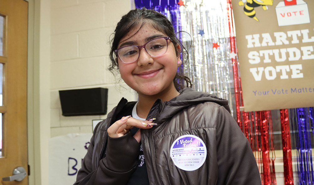 Hartley Elementary School fifth grader Haniya proudly displays an "I Voted Today" sticker on her jacket while standing in a school hallway. She is standing in front of a sign that says Hartley Student Vote - Your Vote Matters.