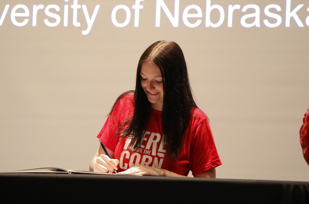 A Northeast High School senior smiles as she prepares to sign a book during a signing ceremony for future educators. She is sitting at a table on stage in Northeast's auditorium and using a pen in her right hand.