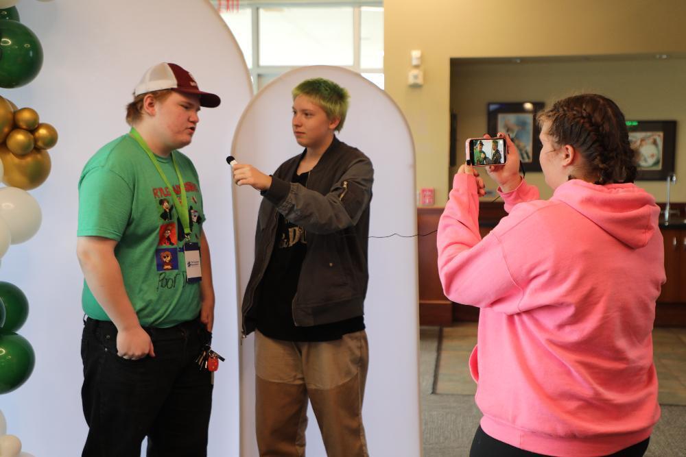 Three Bay High Focus Program students take part in an activity at the Class Intercom Content Generation Workshop this fall. One boy is being interviewed by another boy with a small handheld microphone that is connected to a smartphone by a long wire. A girl is video-recording the interview with the smartphone on the righthand side of the picture.