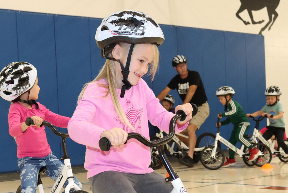 A Maxey Elementary School kindergarten student smiles as she enjoys riding a new Strider bike in the school gym. A physical education teacher and four other students are riding on bikes behind her.