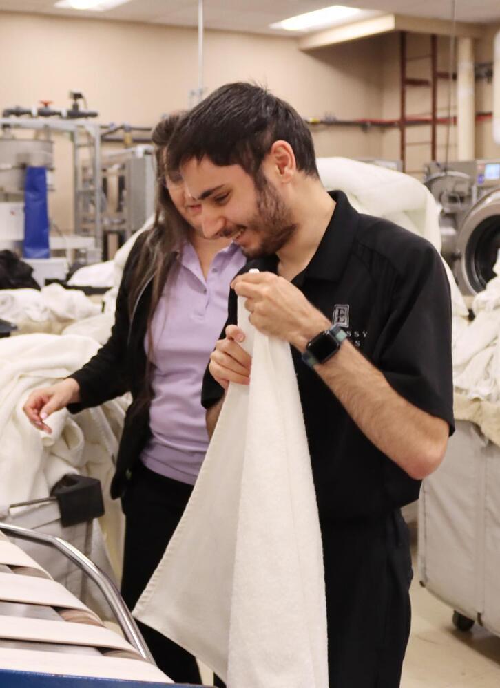 Independence Academy senior Ali folds a towel in a hotel laundry room as a woman stands behind him.
