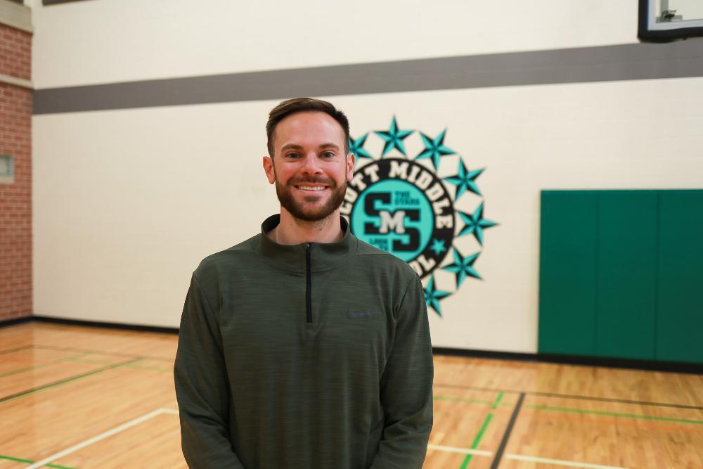 A smiling man with a beard, wearing a dark green quarter-zip, stands in a gym. Behind him is a "Scott Middle The Storm" logo with teal stars on a white wall, above a light wood basketball court.
