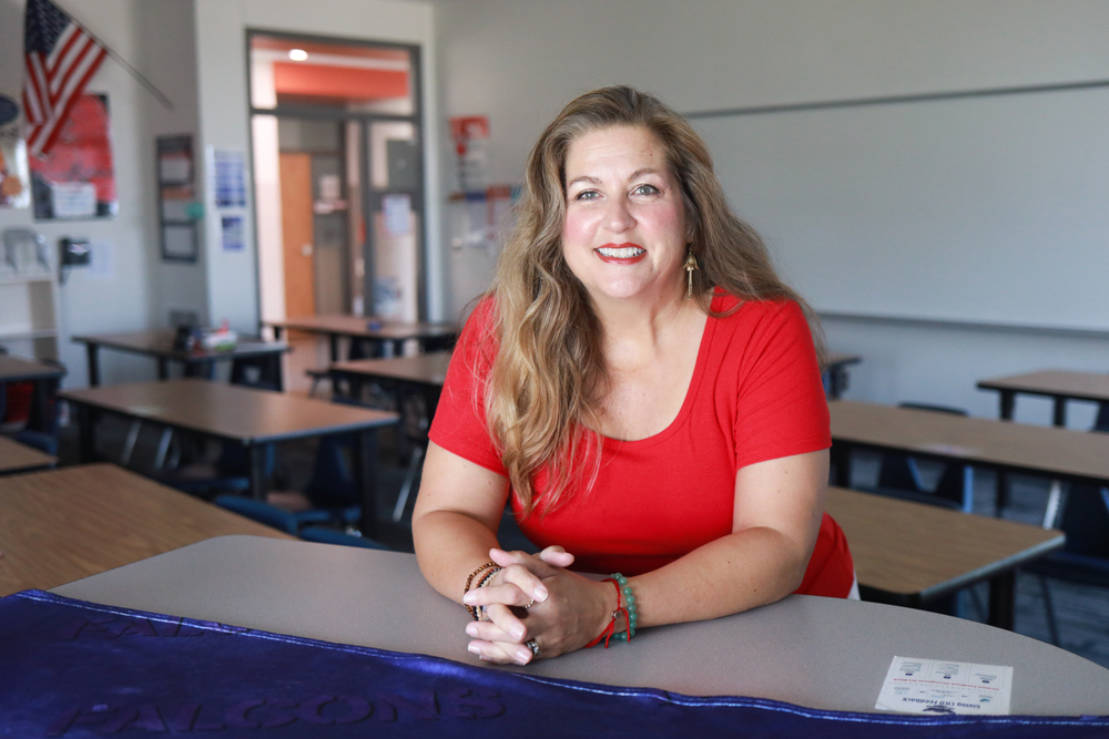 Woman wearing a red shirt smiles in a classroom with her hands together.