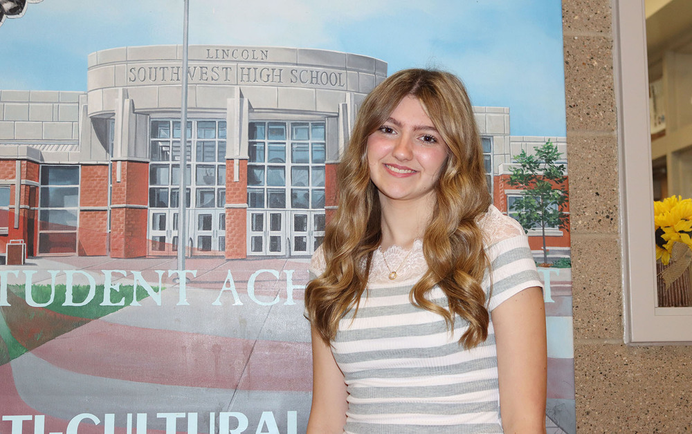 Southwest High School junior Kayleigh smiles in front of a mural that shows the front entrance of the school building. There is a small amount of tan wall that is on the righthand side of the photo. She is the editor-in-chief of the school newspaper.