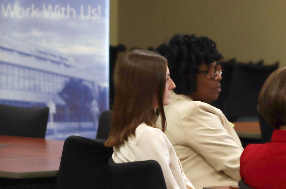 LPS student teacher Sarah Baillie and LPS Associate Superintendent for Human Resources Vann Price listen to a group of teachers and administrators at a student teacher event this fall. There is a banner behind them that says Work With Us! and they are sitting in chairs at a rectangular table.