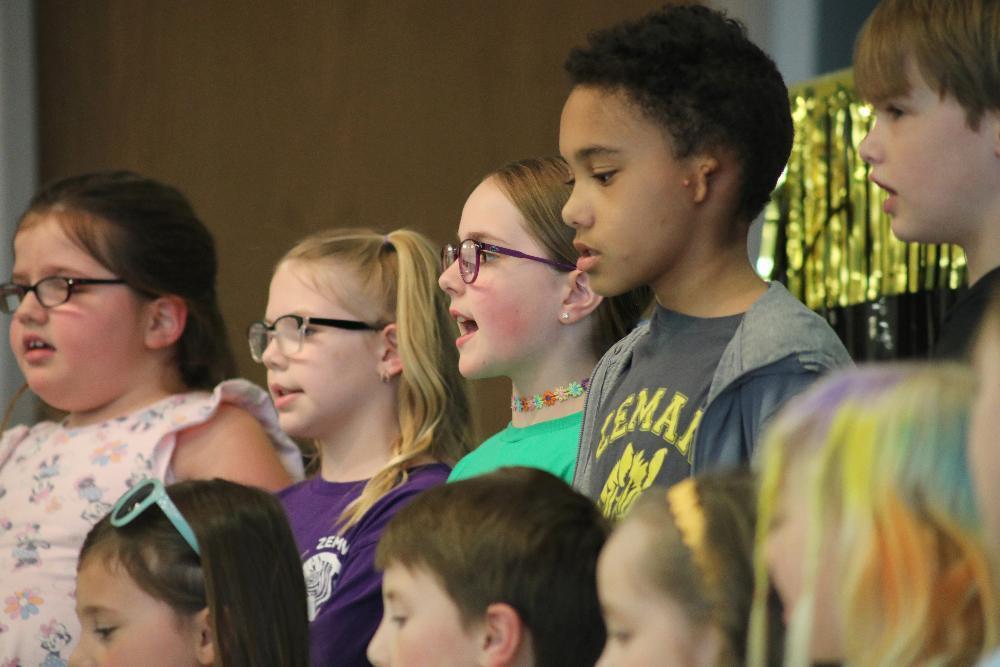 Children in glasses and casual clothing sing in a group. One boy wears a grey sweatshirt with "LEMAI" on it. They are in an indoor setting.