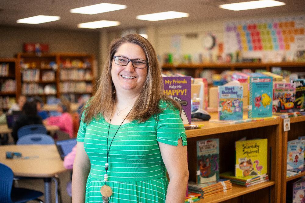 A woman stands in a library, smiling. She wears a green striped dress. Behind her are shelves filled with children's books. One book cover reads, "My Friends Make Me HAPPY!"