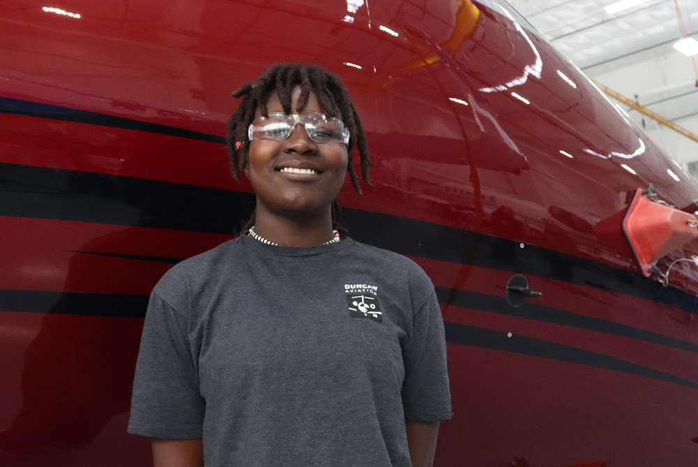 Aviation and Technical Education Focus Program senior Elizabeth stands in front of an airplane. She is wearing safety goggles and a t-shirt that says Duncan Aviation on it.