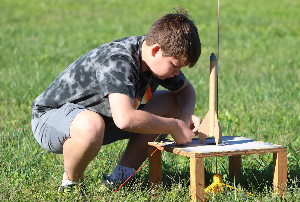 A Mickle Middle School student is crouching down next to his rocket that is ready to launch on a wooden four-legged platform. He is connecting wires to the base of the rocket to allow him to press the ignite button while safely standing away from the platform.