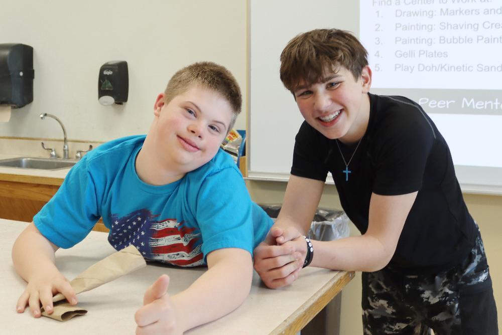 Two boys work together at a table in a classroom. One boy is crafting with paper; the other assists. The classroom includes a sink and whiteboard listing art supplies.