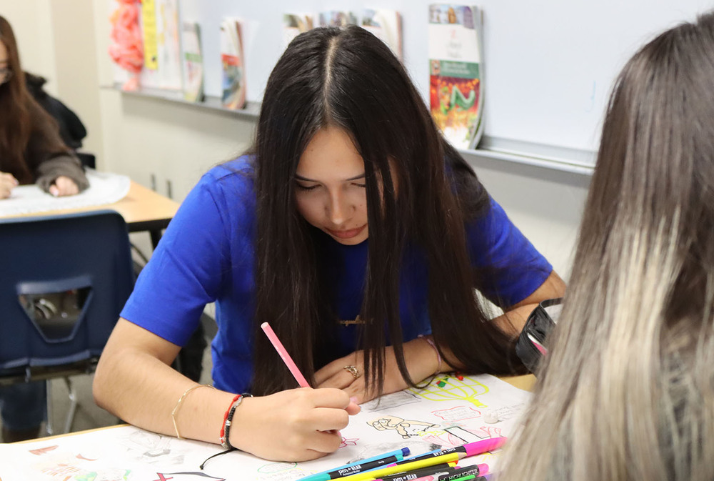 A North Star High School student concentrates as she draws on her mural during her Spanish for Spanish Speakers Level 3 class. She is holding a pencil with her right hand and is sitting at a classroom table.