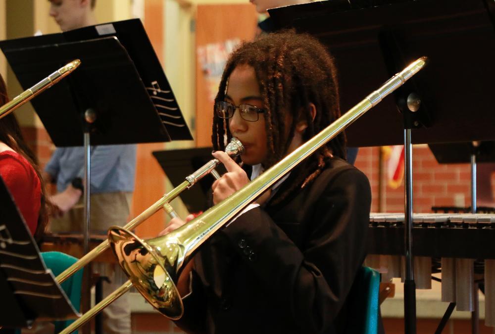 A Lux Elementary School student is playing a trombone in the school band during a Veterans Day assembly. He has glasses and braided hair.