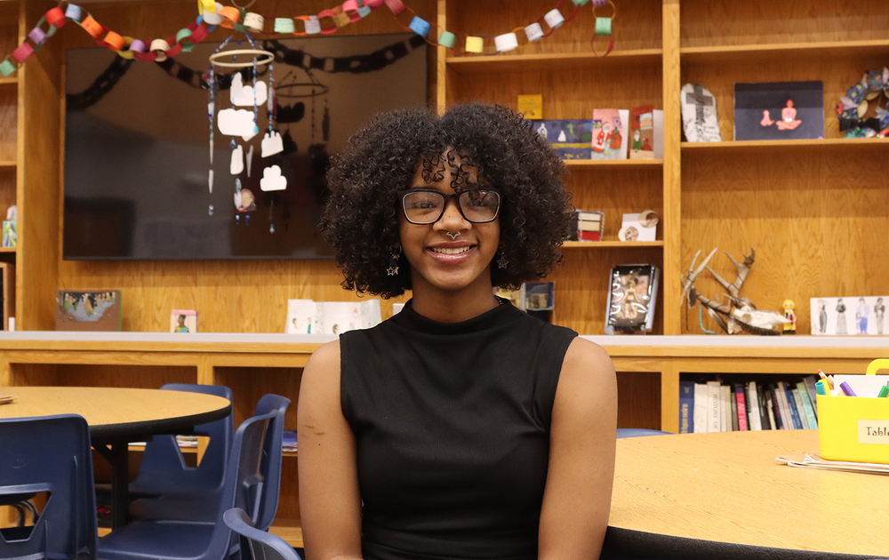 Arts and Humanities Focus Program senior Alainah sits at a table in the school's commons area. A television screen and several rows of bookshelves are behind her.