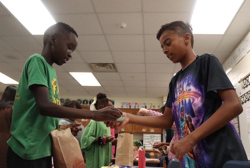 Two boys exchange a light blue object in a brightly lit classroom. The boy in a green shirt hands it to the boy in a purple Space Jam shirt. Other children are visible in the background, participating in activities around brown paper bags on tables.