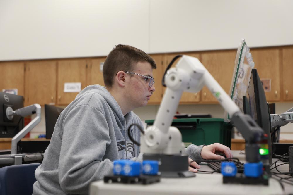 A student in glasses and a grey hoodie focuses on a computer monitor, his hands on a keyboard. A white robotic arm is prominent on the desk, with blue blocks nearby. Wooden cabinets line the background, suggesting a classroom or lab setting.