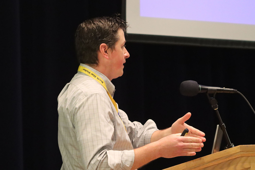Nebraska Public Power District employee Taylor Sutton speaks to North Star High School students about Cooper Nuclear Station. He is standing at a wooden podium with a large microphone in front of him. There is a video screen hanging down behind him.