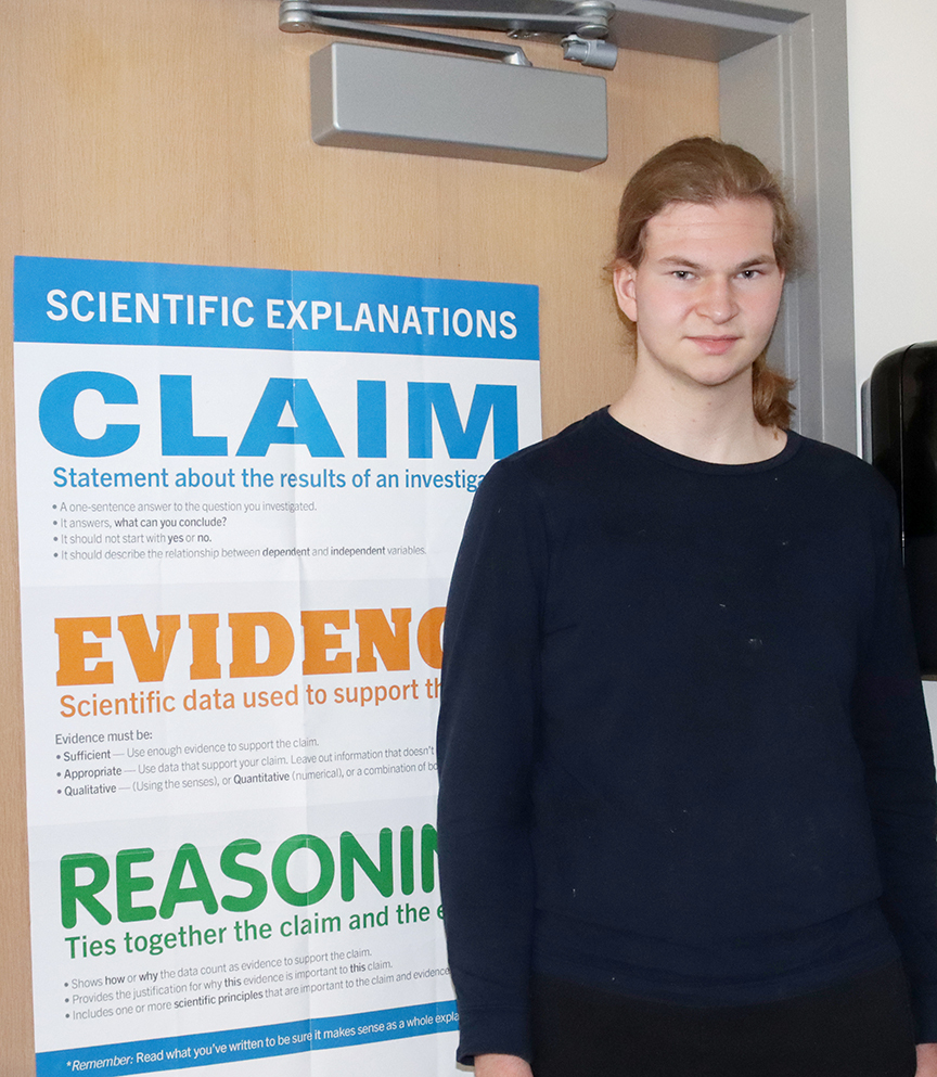 Science Focus Program senior Malcolm smiles in front of a door that has a poster on it. The poster has the title of Scientific Explanations and the headlines of Claim, Evidence and Reasoning on it.