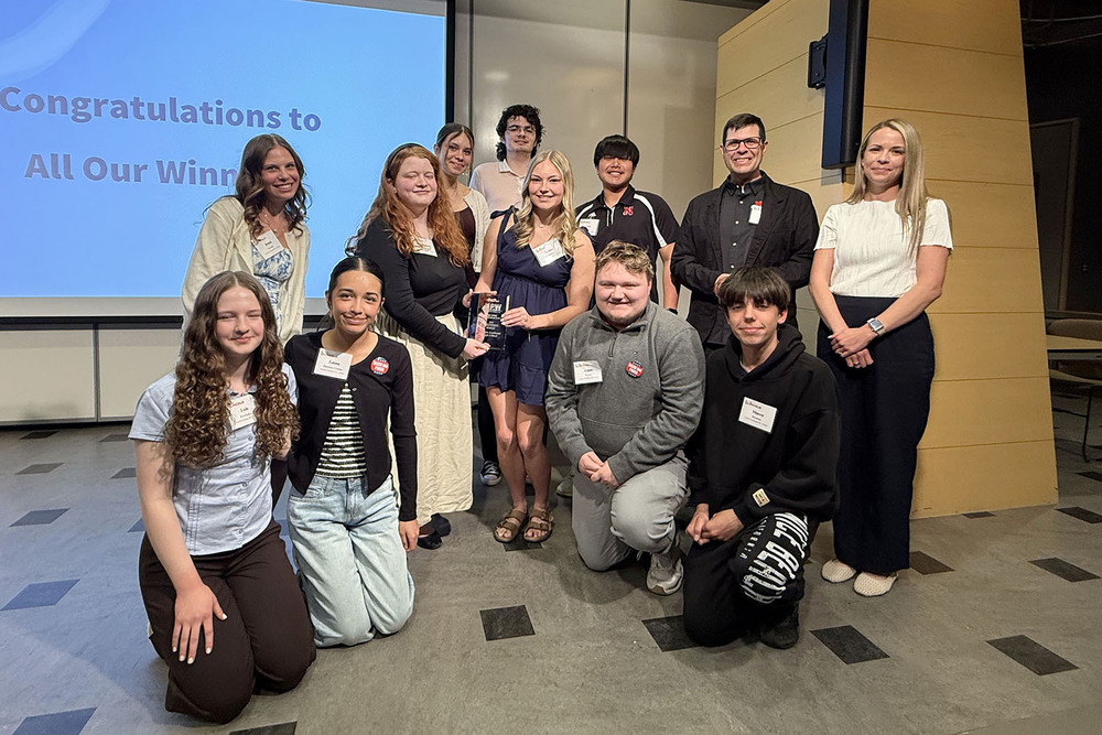 Ten Southeast High School students and two adults smile with their awards at the Nebraska Press Women High School Communications Contest. Two students in the middle of the group are holding a glass plaque the school received for winning the team championship with 19 awards. Four students are kneeling down and the remaining eight people are standing.